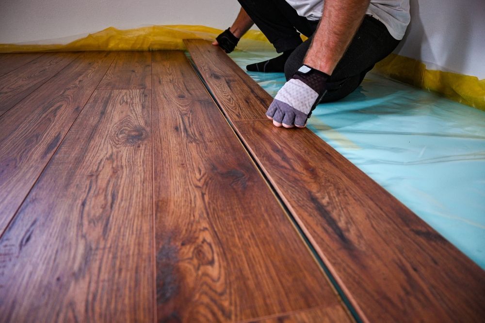 Person installing laminate flooring, kneeling, placing a plank. Brown wood, blue moisture barrier.