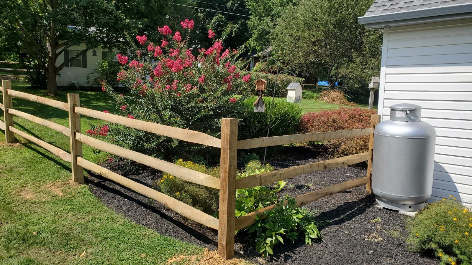 Wooden split-rail fence bordering a garden with flowering bushes, and a propane tank next to a white wall.