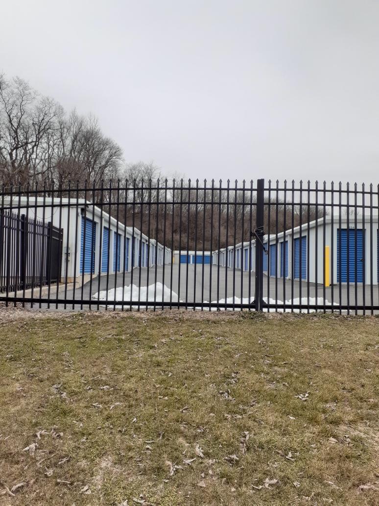 Storage units behind a black fence. Units are white with blue doors, set in grassy area under overcast sky.