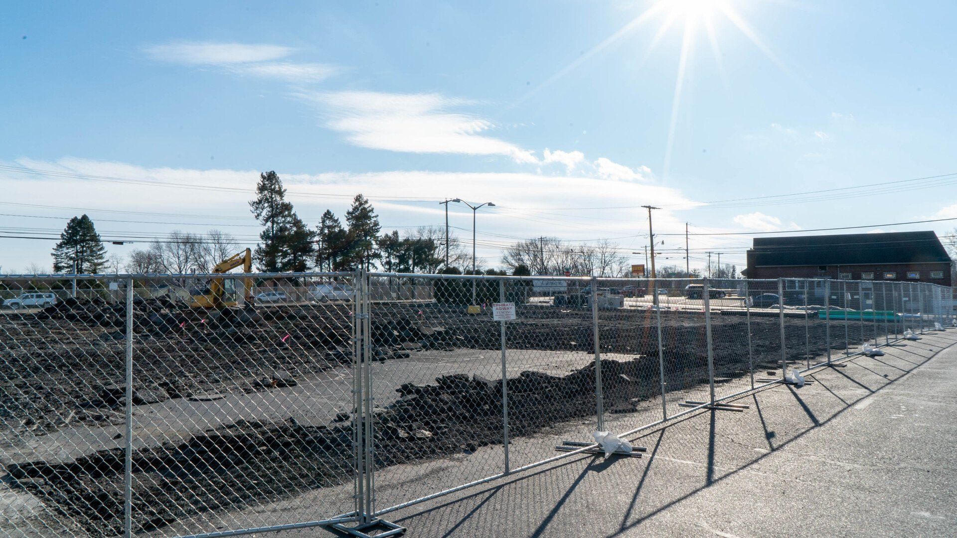Construction site behind a chain-link fence on a sunny day. A small building is visible in the background.