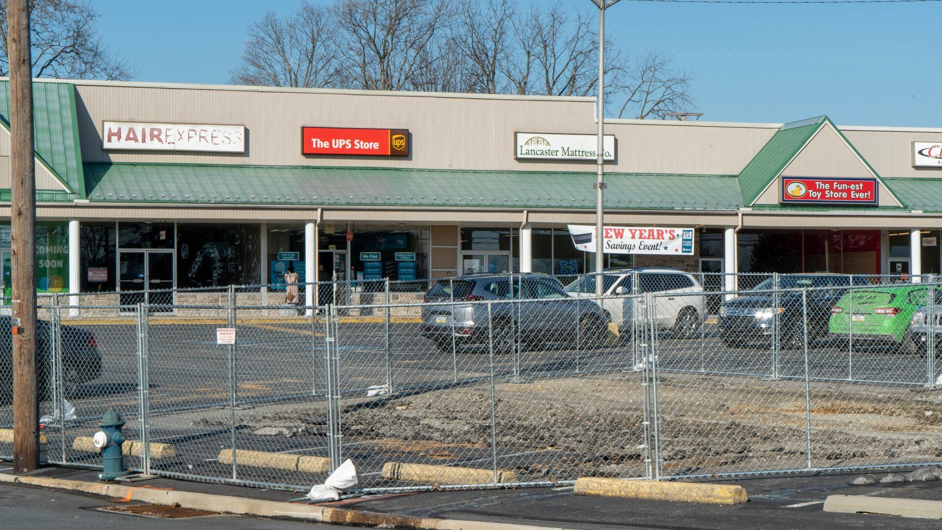 Shopping center with several businesses, parking lot, and chain-link fence. Cars parked in front.