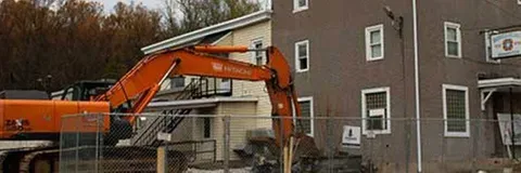 An orange excavator demolishes a two-story building, likely for construction. Trees and a fence are in the background.