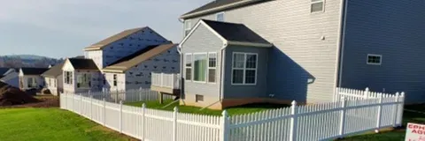 White picket fence surrounds a green lawn in front of gray houses on a sunny day.