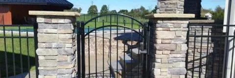 Stone pillars with a black iron gate. Green grass and trees in the background, under a blue sky.