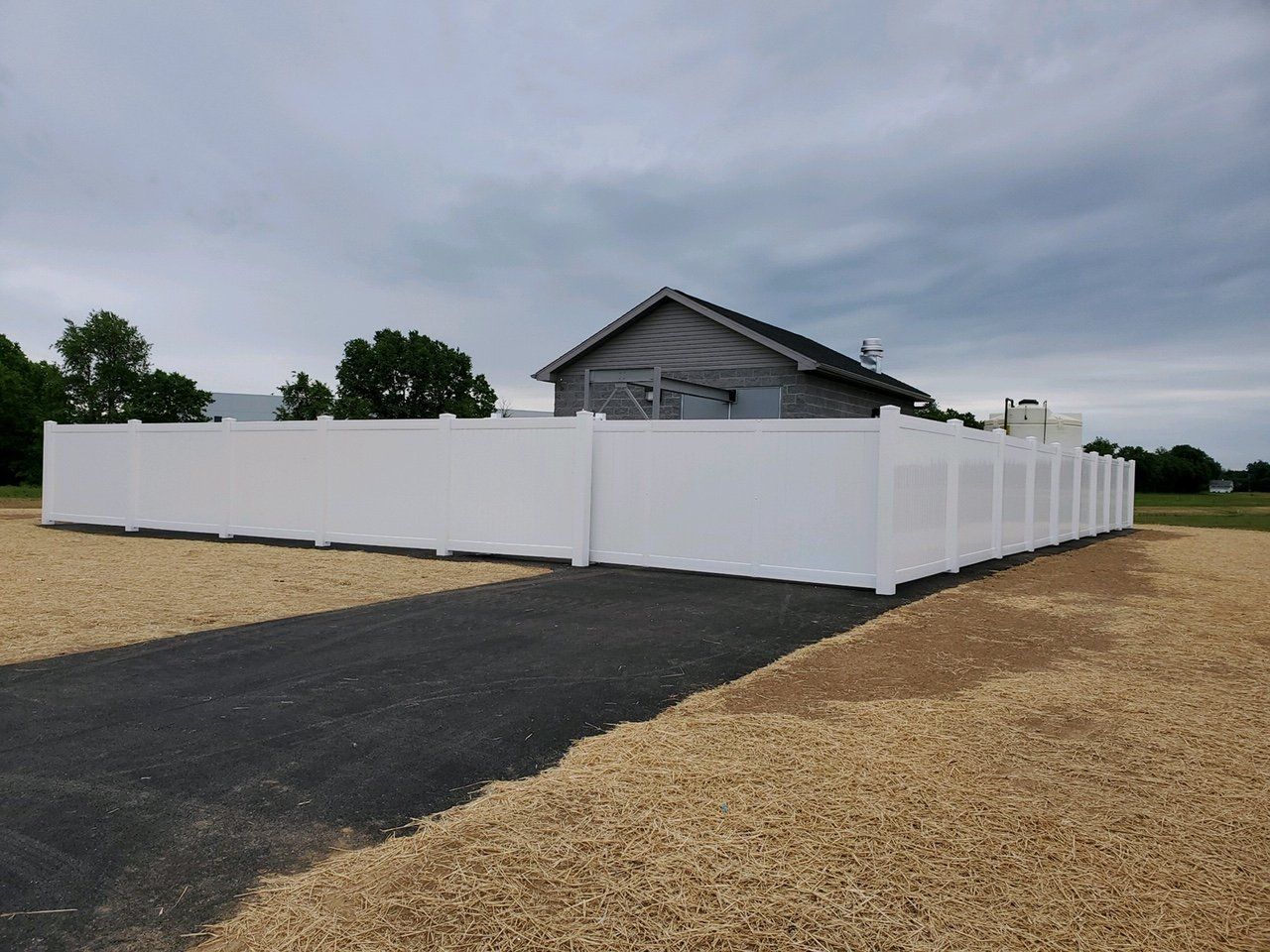 White fence surrounds a gray building on a gravel lot with a paved driveway under a cloudy sky.