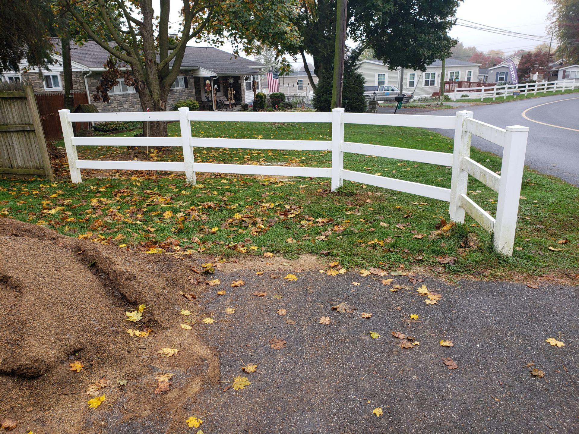 White fence curving along a road, with a pile of mulch in front and houses in the background.