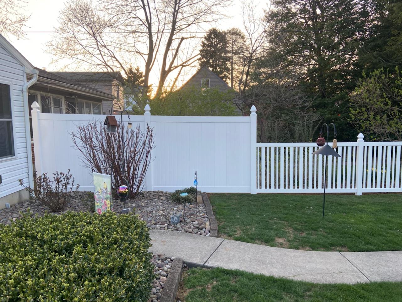 White picket fence surrounds a lawn with a sidewalk leading to a house; trees and sunset in the background.