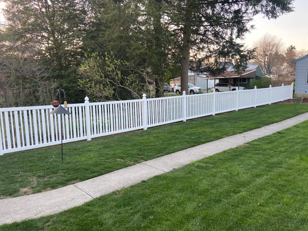 White picket fence bordering a green lawn with a sidewalk. Trees and houses in the background.
