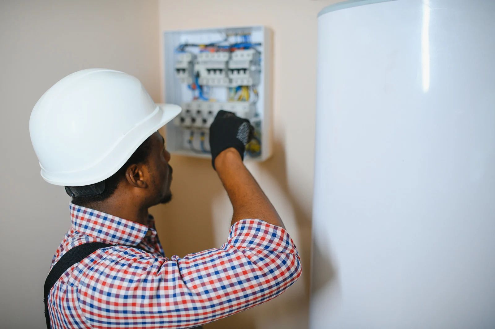 Electrician wearing a hard hat and gloves working on a circuit breaker panel next to a water heater.