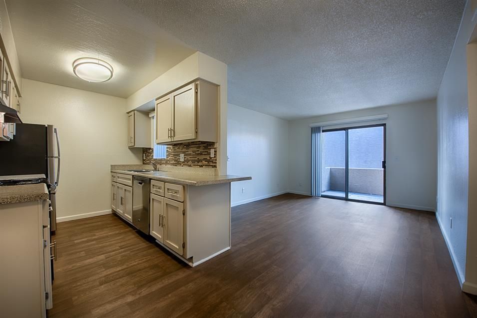 Empty apartment kitchen and living area with wood floors and sliding glass door.