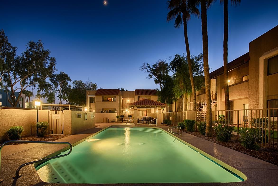 Lit apartment pool glowing green at dusk, flanked by palm trees and buildings.