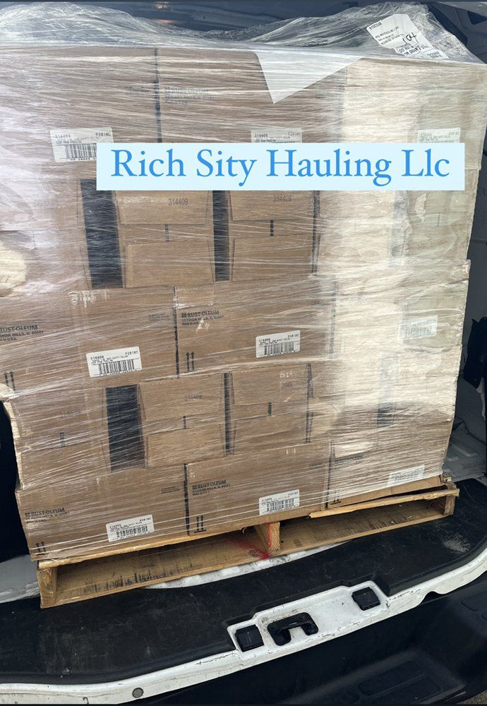 Pallet of boxed goods wrapped in plastic on a truck bed, labeled “Rich Sity Hauling Llc.”