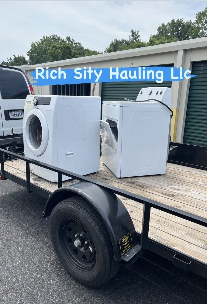 Two damaged white appliances strapped on a flatbed trailer in front of a Rich City Hauling truck.