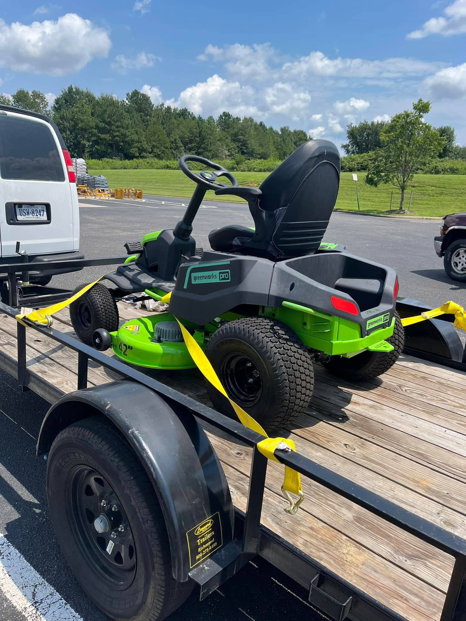 Bright green riding lawn mower strapped to a trailer beside a white truck outdoors.