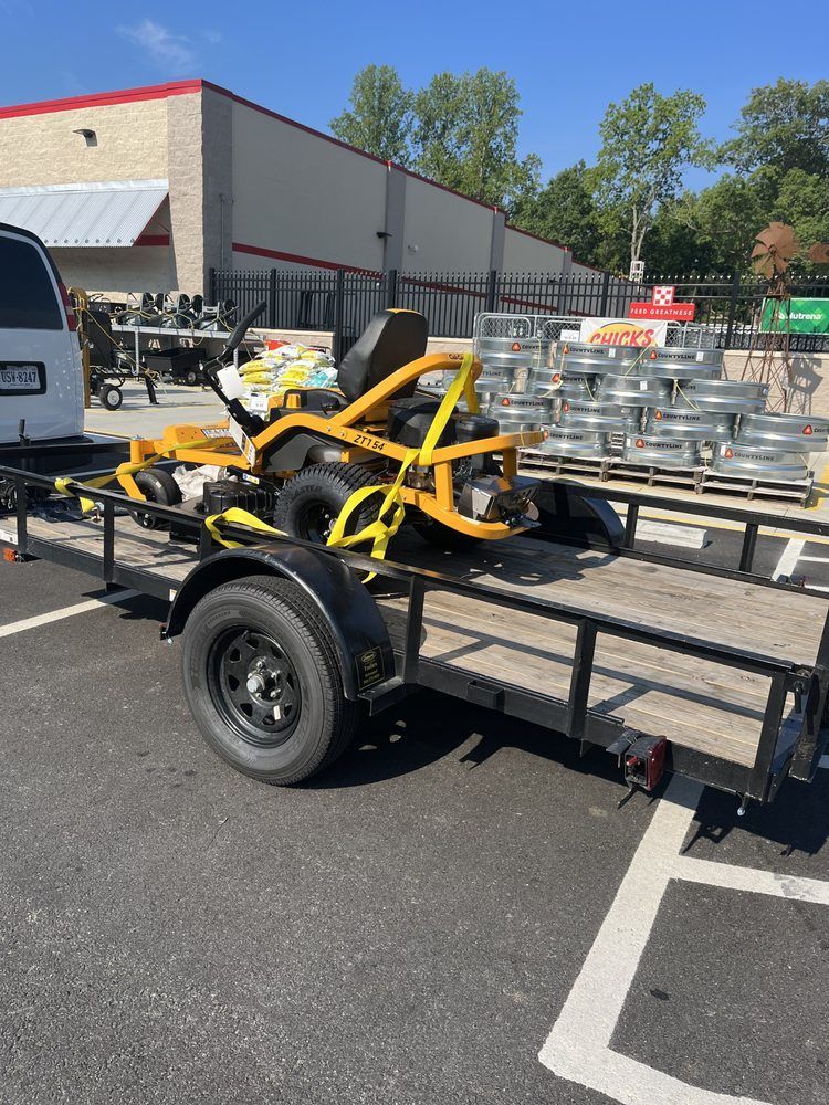 Yellow go-kart on a black trailer in a parking lot near stacked metal pallets and a building.
