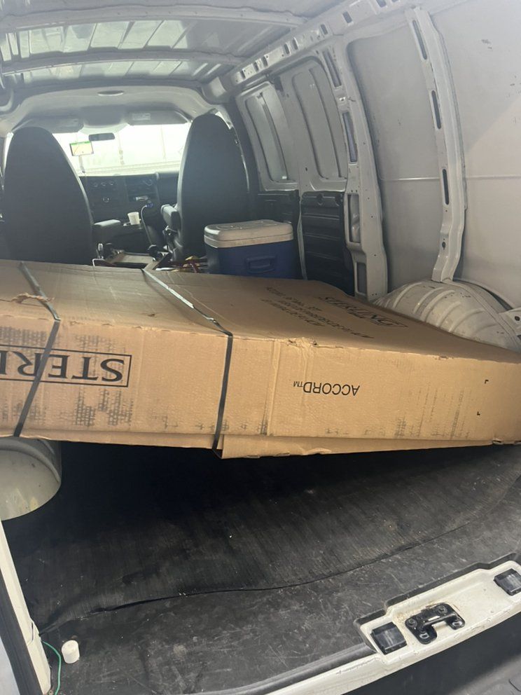 Large cardboard box strapped inside a white cargo van, with a suitcase and tire nearby.