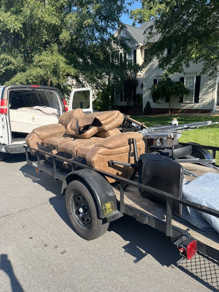 Black utility trailer loaded with tan furniture being towed behind a white van on a suburban street