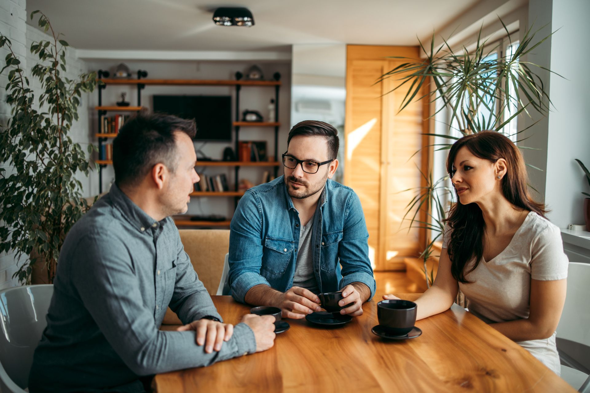 Three people seated at a wooden table, conversing. Indoors, bright room with a TV and plants in the background.