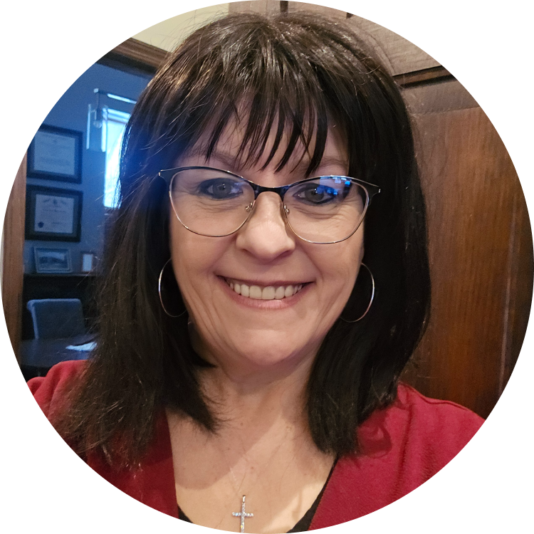 Woman with glasses smiles; dark hair, red top; office setting with artwork in background.