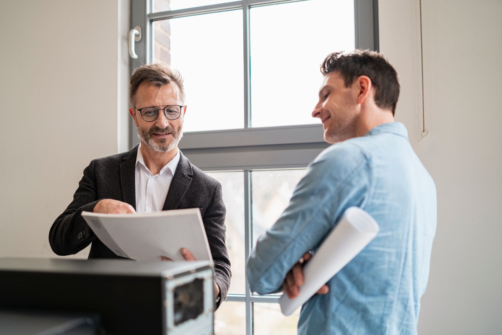 Two men in an office looking at documents near a window. One man smiles, holding rolled paper.