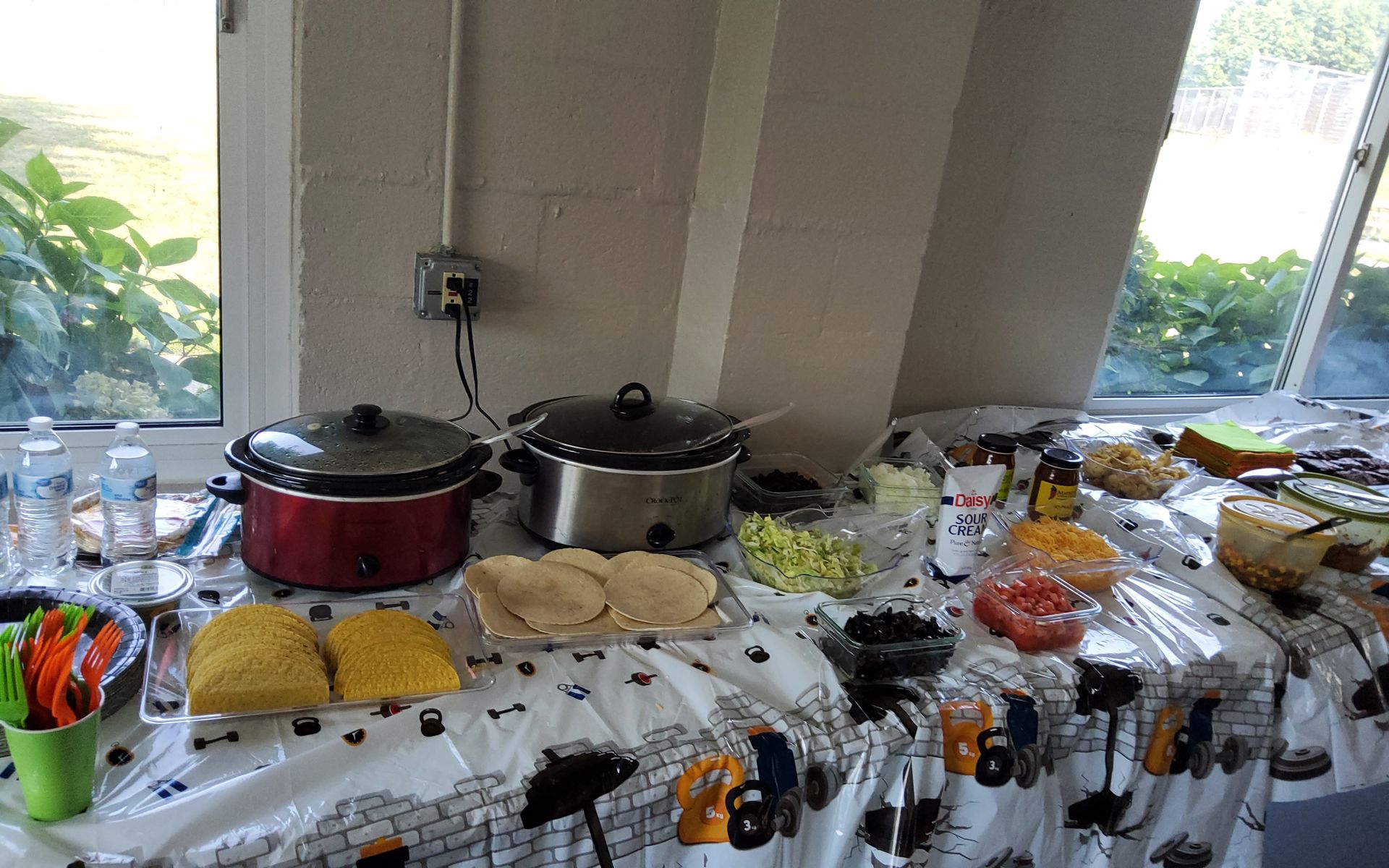 Buffet table with taco fixings: two crockpots, tortillas, toppings, and drinks near a window.