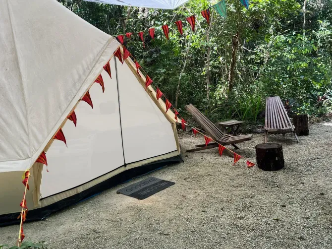 Carpa de lona blanca con banderas rojas, sillas de madera y suelo de grava en una zona boscosa.