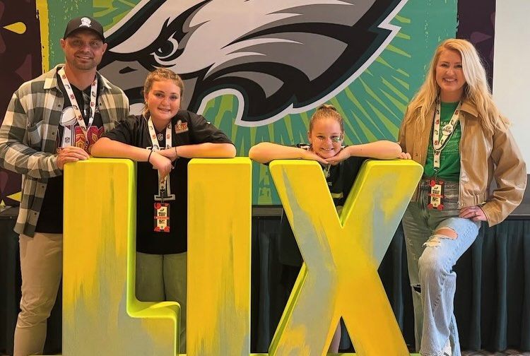 Four people posing behind large yellow letters LIX in front of an Eagles mural