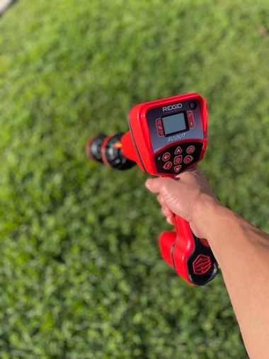 Hand holding a red handheld device over green grass, aimed at the ground