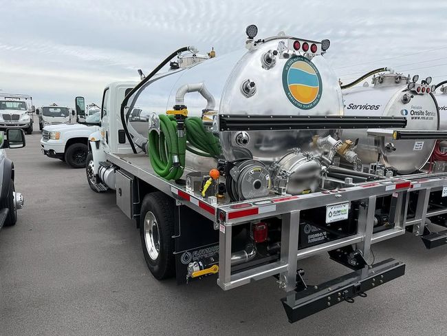 White vacuum truck parked in a lot, with silver tanks, hoses, and rear equipment.