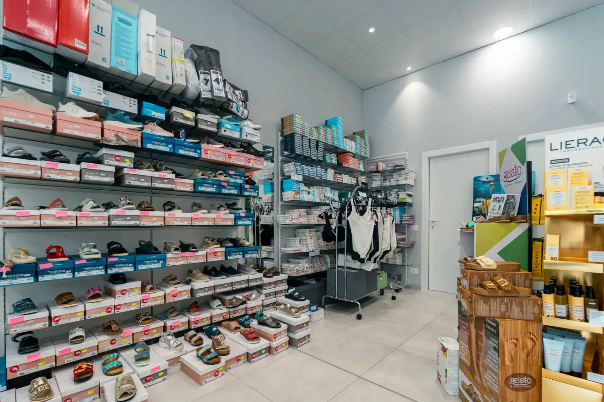 Interno di un negozio di scarpe con scaffali pieni di scarpe colorate, borse e appendiabiti in una stanza bianca e luminosa.