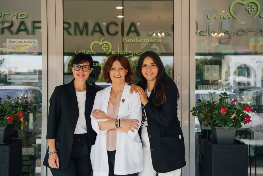 Tre donne posano sorridenti davanti all'ingresso di una farmacia, abbracciate.