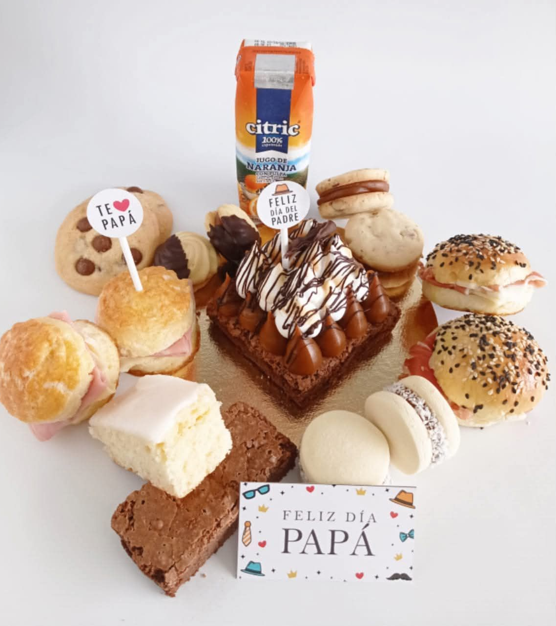 Surtido de postres para el Día del Padre con pasteles, tarta y una tarjeta de “Feliz Día Papá” sobre fondo blanco.