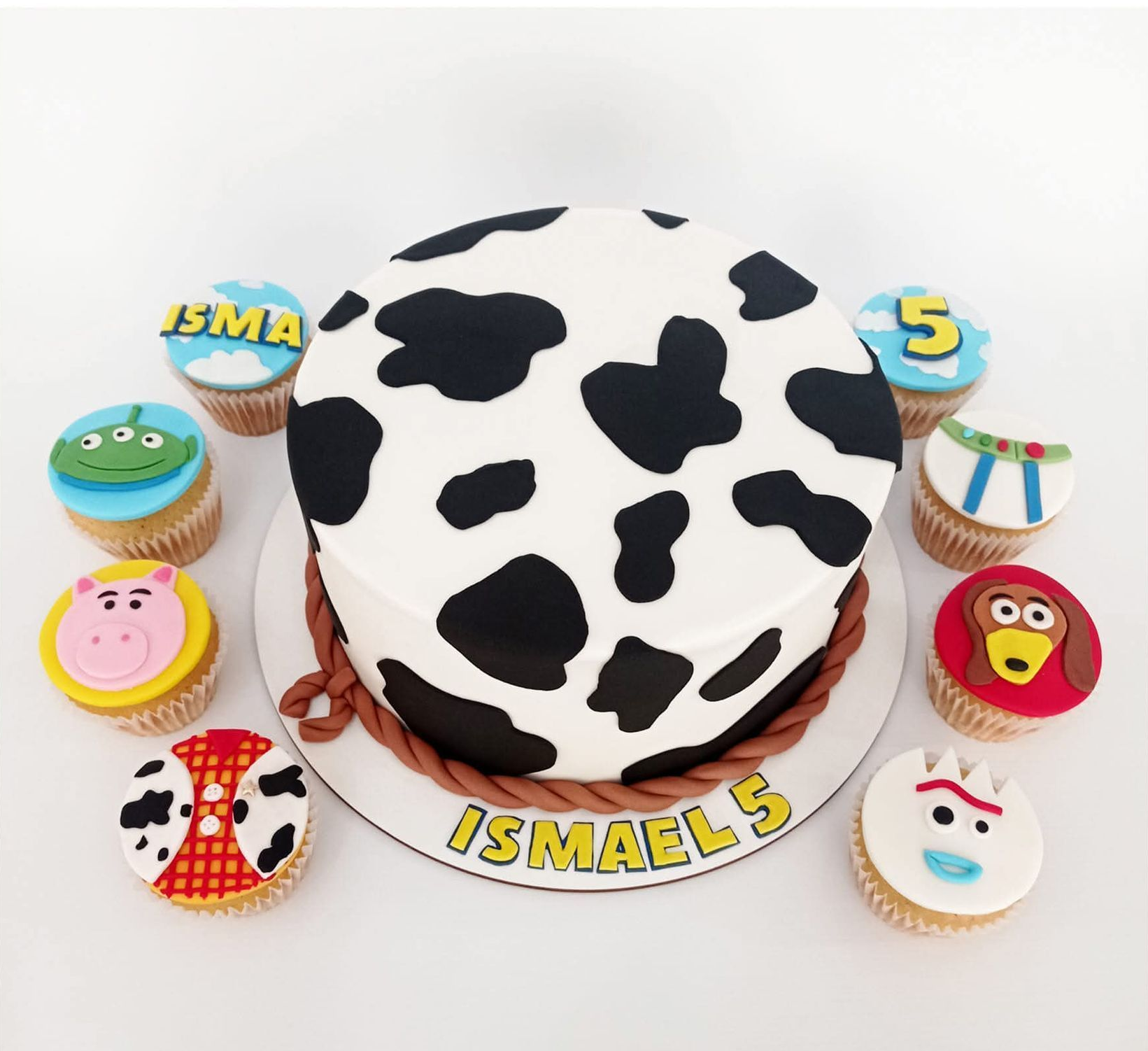 Tarta de cumpleaños con estampado de vaca rodeada de coloridos cupcakes decorados con el nombre Ismael en la tabla.