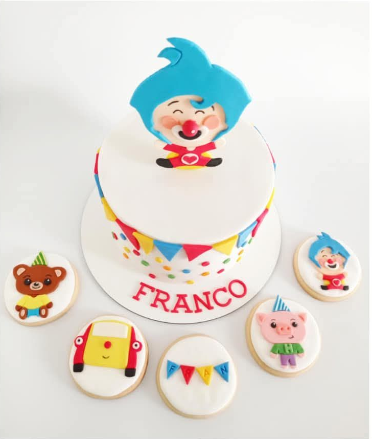Pastel blanco con adorno de dibujos animados azul, el nombre “FRANCO” y galletas de personajes.