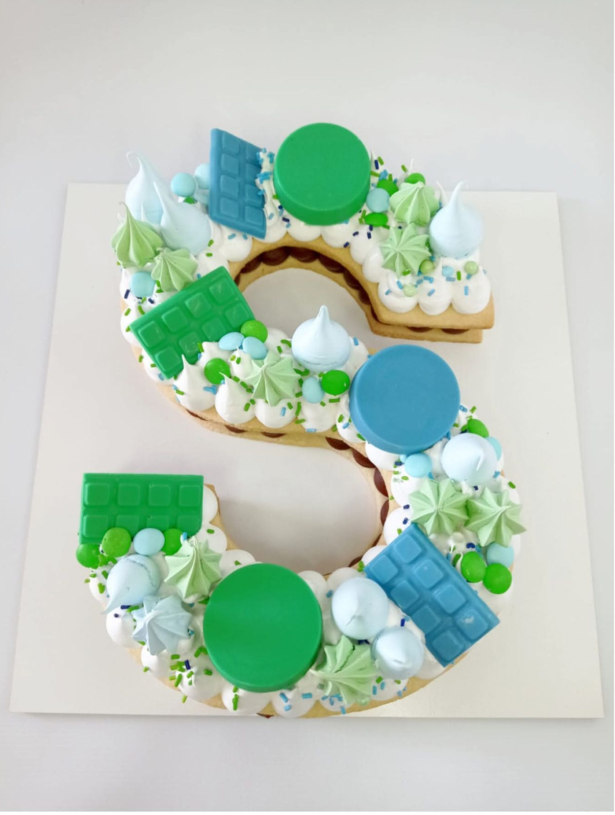 Tarta decorada con el número 5, con glaseado verde, azul y blanco y adornos redondos sobre una base blanca.