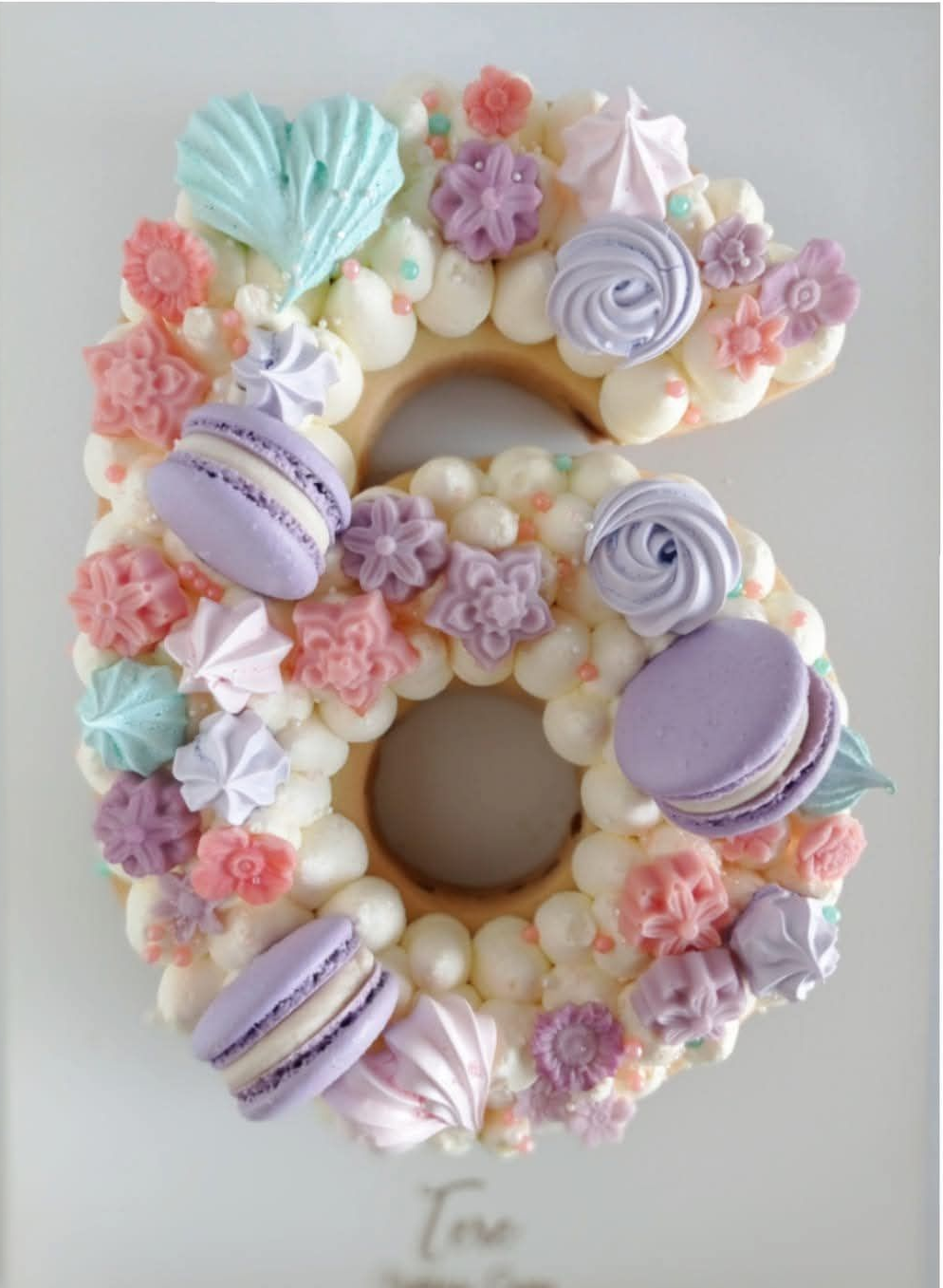 Tarta número 6 de macarons en tonos pastel con flores y espirales de crema sobre fondo blanco.