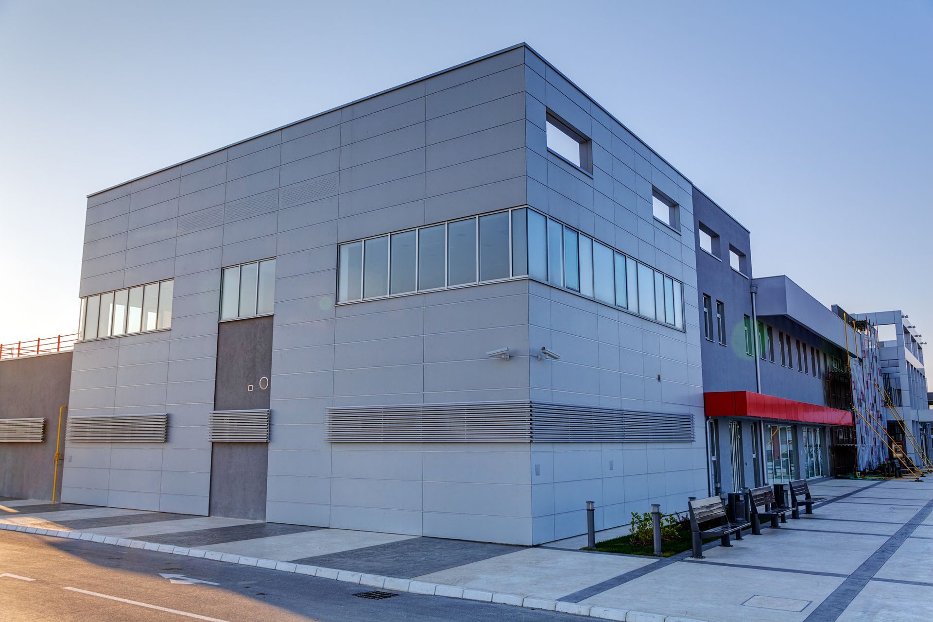 Moderno edificio angolare grigio con ampie finestre su una strada tranquilla, tenda da sole rossa e fioriere sul marciapiede.