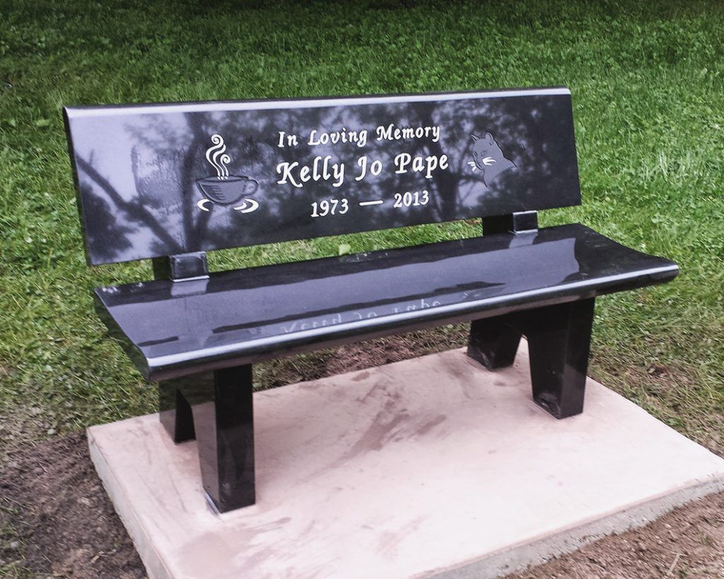 Black memorial bench with engraved tribute, set on a concrete pad in grass