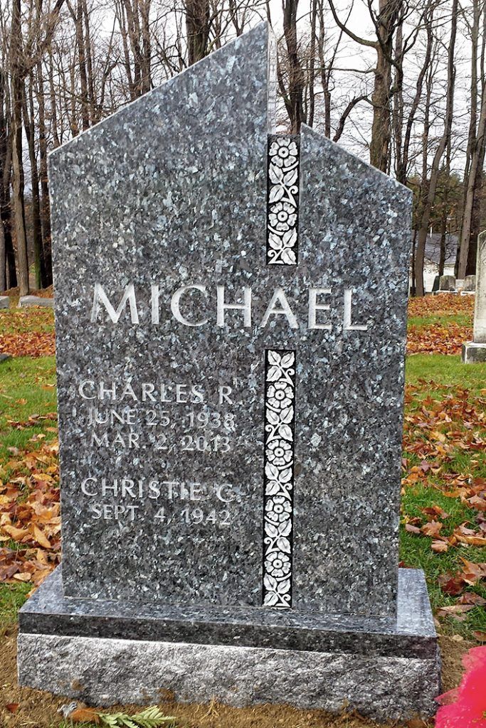 Gray gravestone with the name MICHAEL in a wooded cemetery, with fallen leaves and a red flower at the base