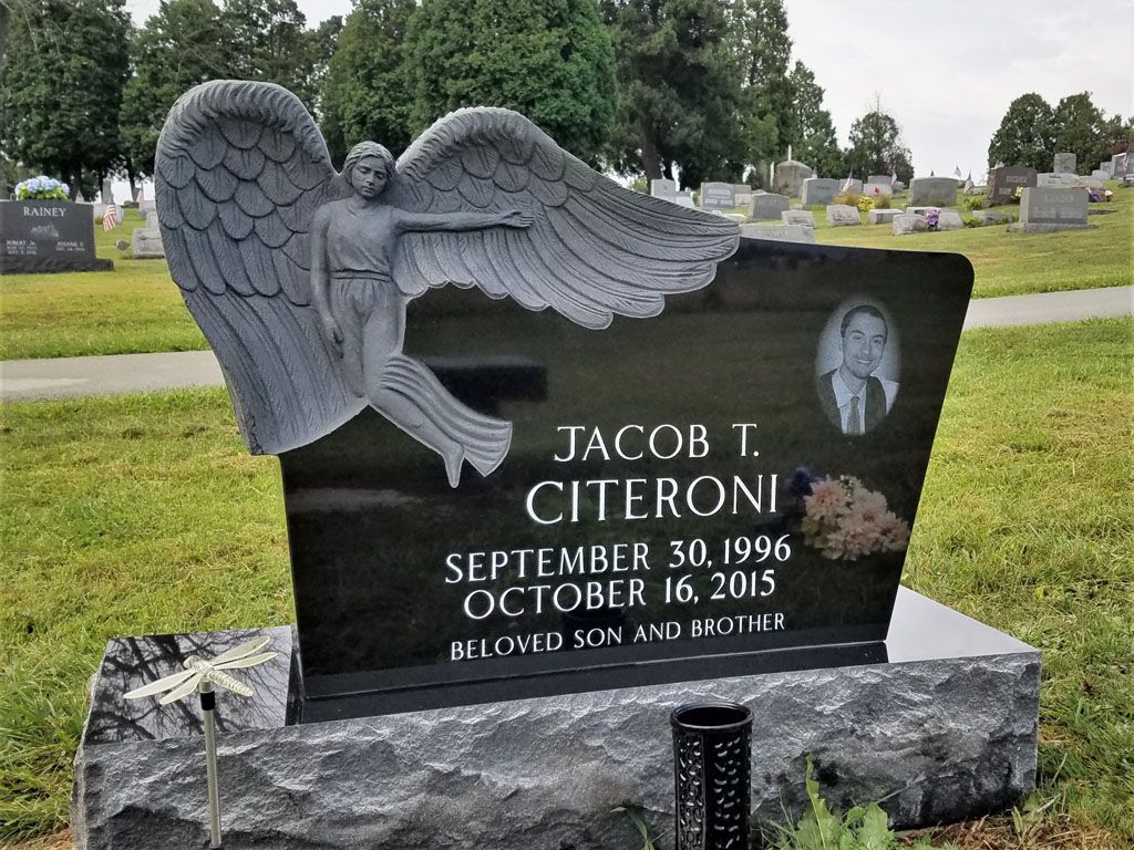 Black cemetery headstone with winged angel relief and an oval portrait in a grassy graveyard.