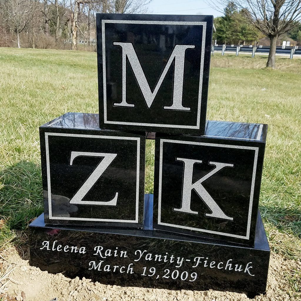 Black granite memorial with stacked letters M, Z, and K in a grassy park; inscription below reads March 19, 2009