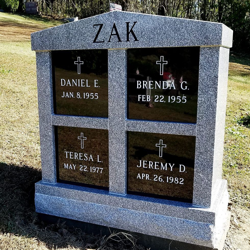 Gray granite memorial with four engraved plaques and crosses, labeled ZAK, standing outdoors on grass.