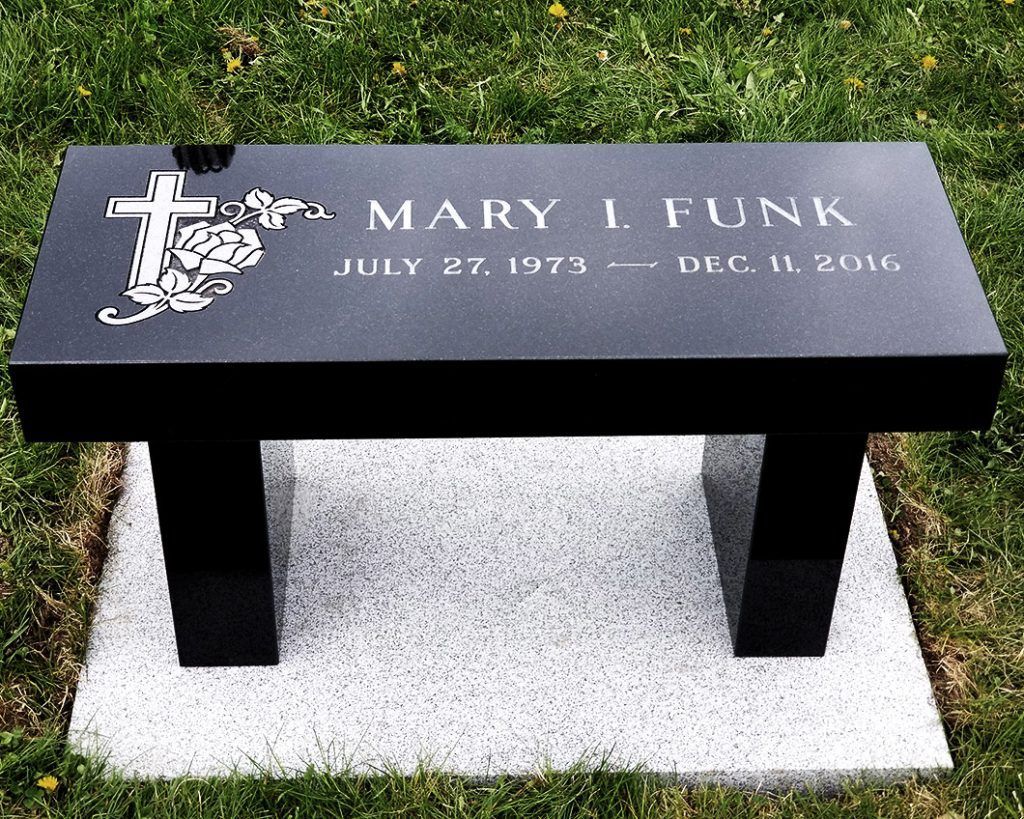 Black memorial bench gravestone with white engraving, set on a light stone base in grass.