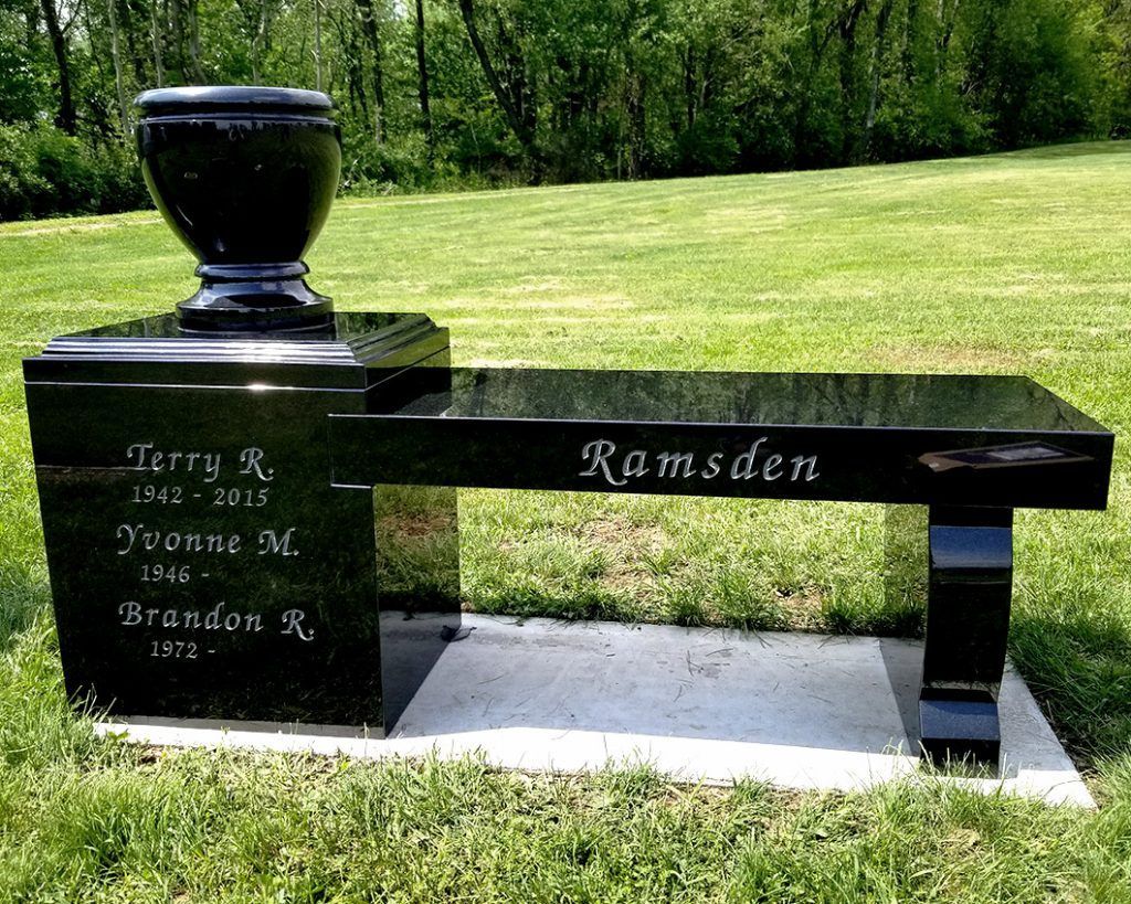 Black memorial bench and urn-shaped monument on a grassy lawn, engraved “Ramsden.”