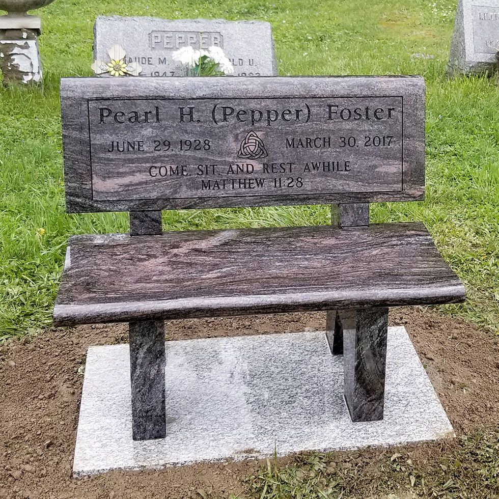 Wooden bench memorial beside a gravestone in a grassy cemetery.