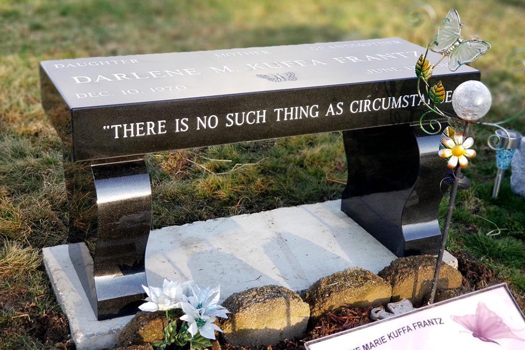 Memorial bench engraved “THERE IS NO SUCH THING AS CIRCUMST...” with flowers and small stones nearby