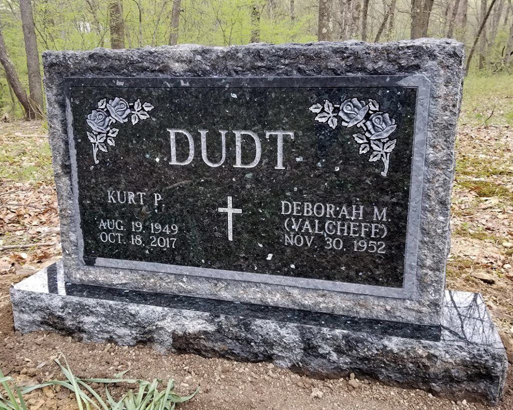 Black granite grave marker with “DUDT” engraved, set in a wooded cemetery.