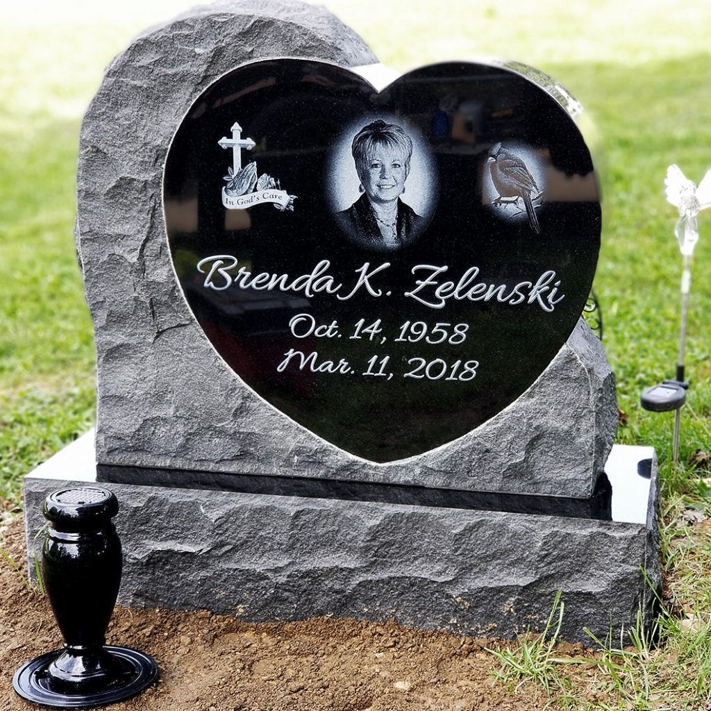 Heart-shaped gravestone with portrait and inscription in a cemetery, beside a black vase and white lamp.