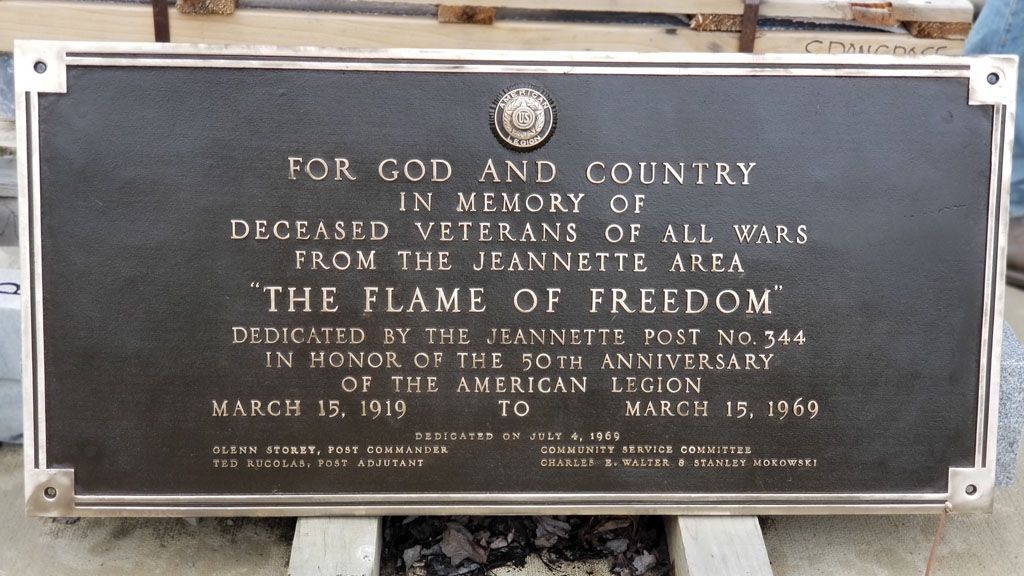 Plaque honoring deceased veterans, titled “The Flame of Freedom,” with bronze text on a dark stone marker.