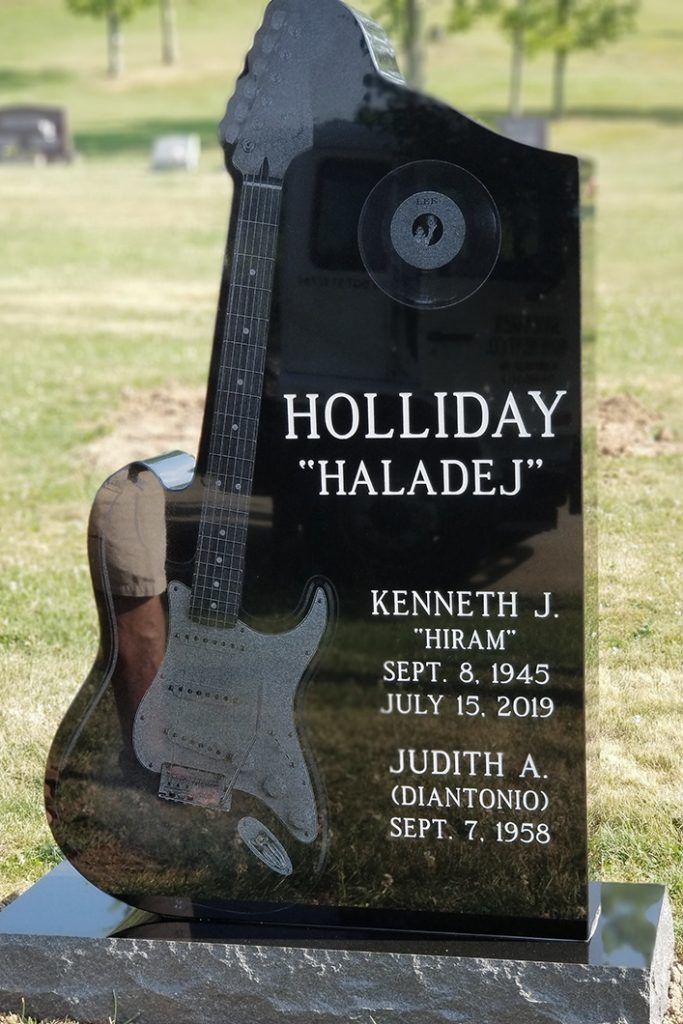Black granite memorial headstone with engraved guitar and the names Holliday “Haladel” and Kenneth J. Holaday.